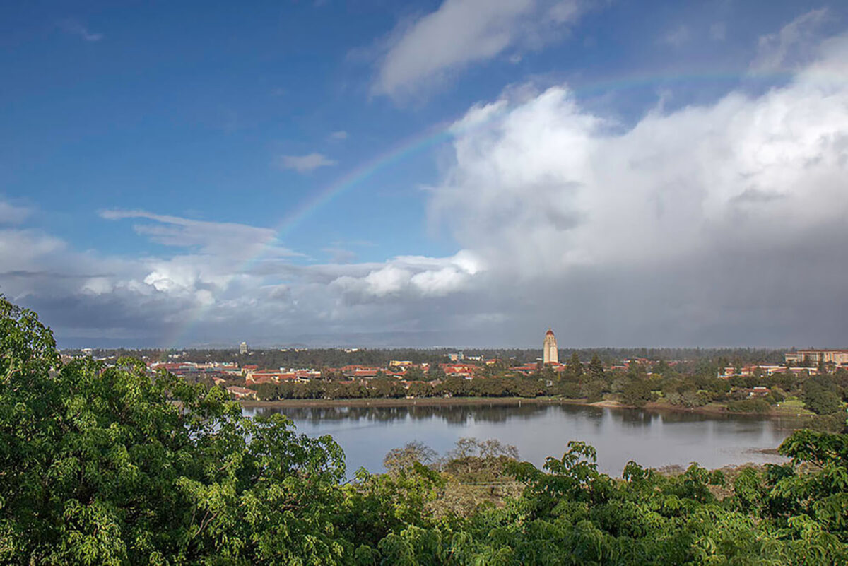 Recent color photograph of Lake Lagunita after winter rain, with water in the basin, Stanford campus buildings in the background, and a rainbow above the scene.